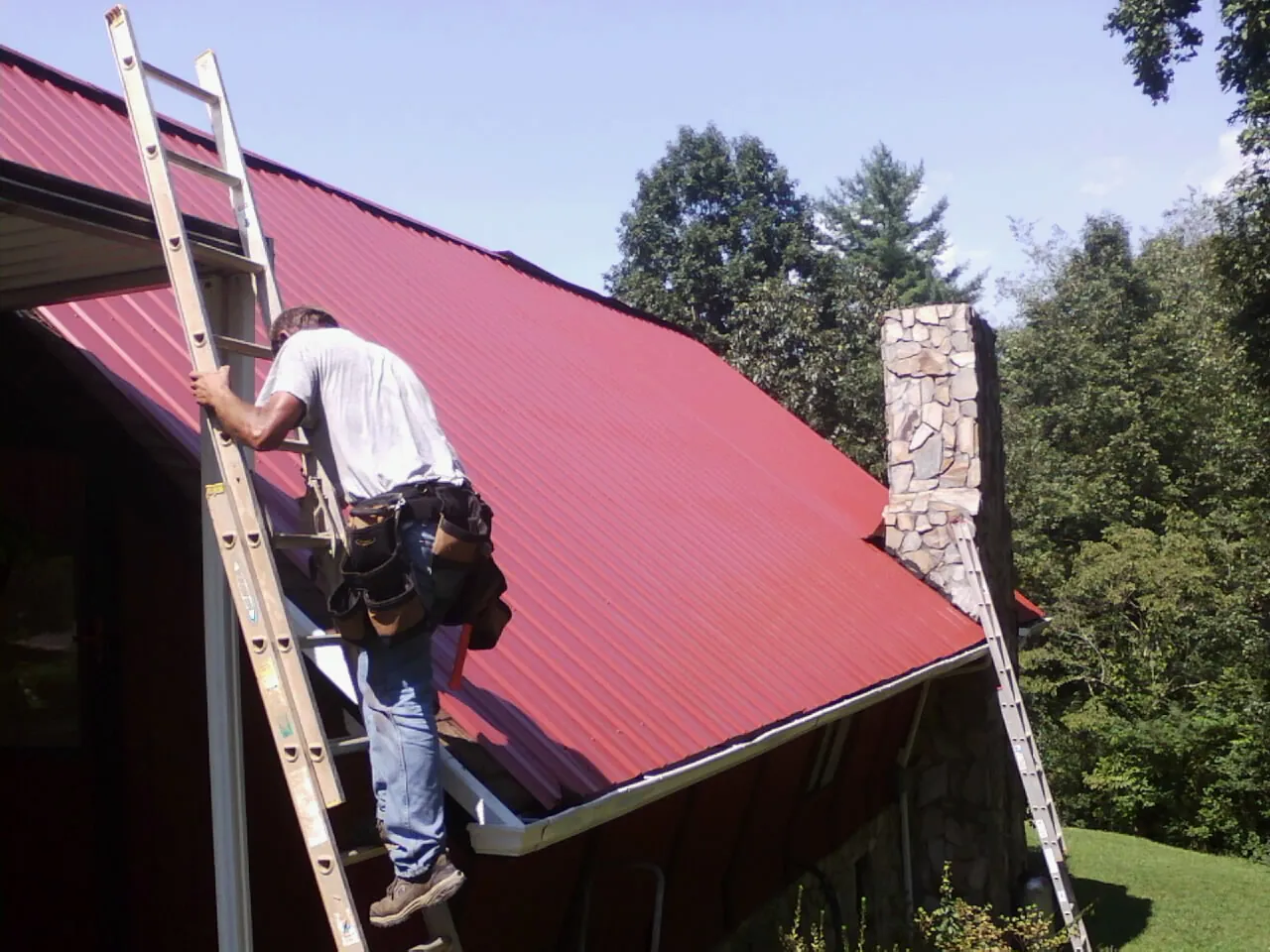 Roofer on a ladder beside a red metal roof and stone chimney — Soffit & Fascia Repair by Roofing Brothers in Rice